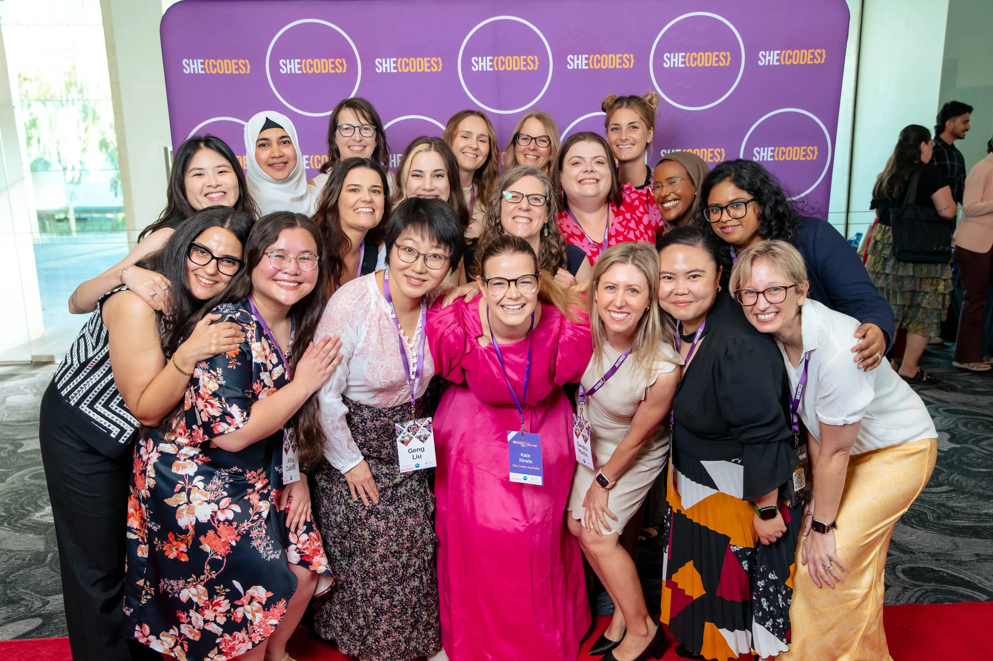She Codes Plus Perth cohort gather for a group hug at Showcase. Kate is in a pink dress in the middle of the group, with the cohort all around her.