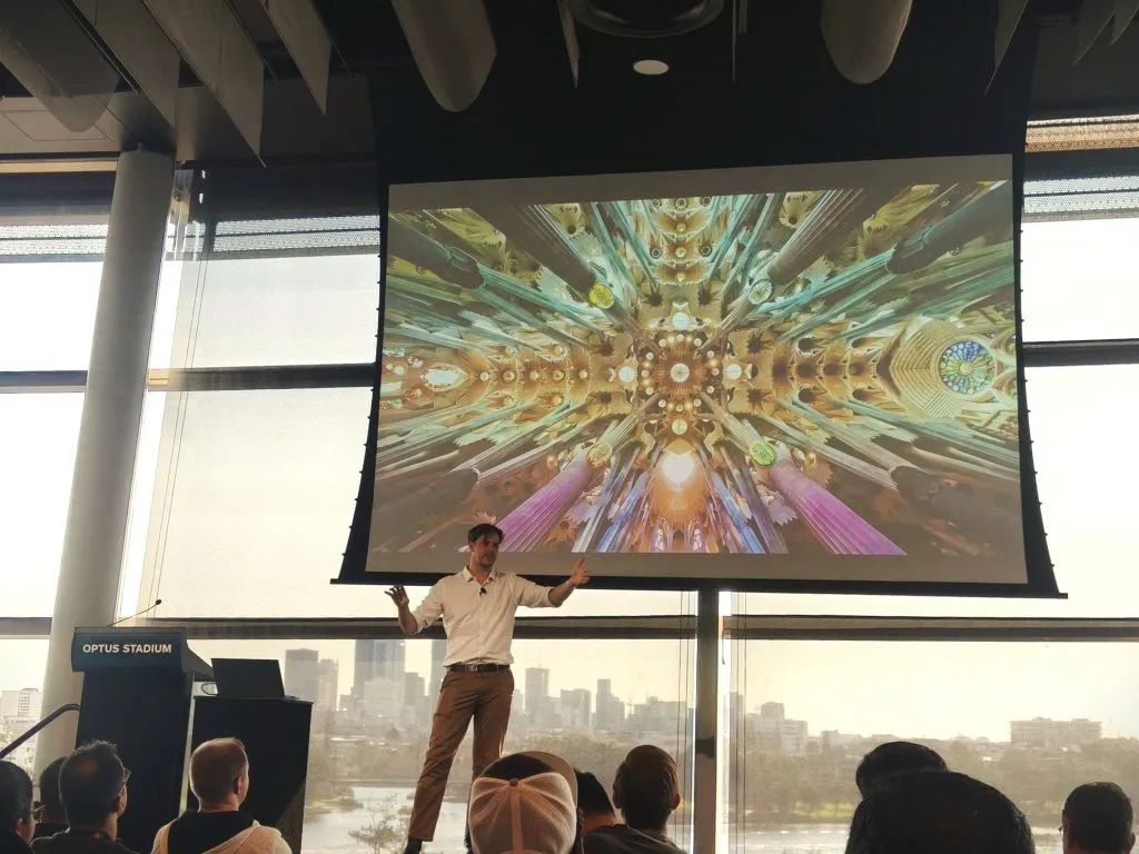 David Smith presents at DDD Perth on automation. He is standing in front of a large screen with a colourful fractal image.